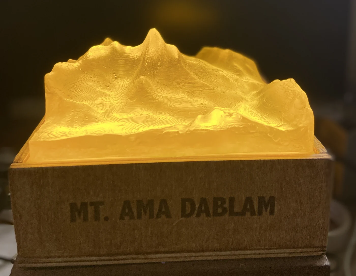 An Ama Dablam terrain lamp switched on in a dark room, the translucent peak glowing warm amber above a wooden base engraved MT. AMA DABLAM.